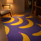 A purple area rug with a bold yellow banana pattern, styled in a warm, modern living room setting next to a wooden chair and bookshelf.