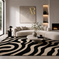 A modern living room featuring a large black and beige wavy pattern area rug under a curved cream sectional sofa. A round coffee table, a black side table with a lamp, a fireplace, and a textured wall art piece with a gold monogram are also visible.