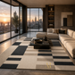 A modern living room featuring a large rectangular area rug with a black, gray, and beige geometric block pattern. The rug is styled in a contemporary living space with a beige sectional sofa, a dark wood coffee table, and large floor-to-ceiling windows showcasing a city skyline at sunset.