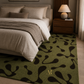 An olive green area rug with a black organic abstract pattern and a small gold monogram, styled in a modern bedroom next to a bed with neutral bedding.