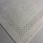 A close-up of a light gray or off-white rug corner showing a geometric pattern of small dark stitched dots forming a V-shape along the edges.