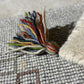 A close-up of a handmade multicolor yarn tassel resting on a woven gray textile surface.