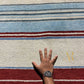 A close-up of a striped rug with cream, light blue, red, and gray horizontal bands, shown with a person's hand and wrist resting on it for scale.