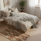 Neatly made bed with beige bedding in a bright bedroom with a plant and rug.