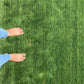 A rectangular green artificial turf or grass mat with a textured grass-like surface, laid out on a light-colored tiled floor. A gold monogram logo reading 'AR' is printed near one corner.