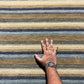 A close-up of a striped area rug with horizontal bands of beige, brown, gray, and muted blue, shown with a person's hand and arm resting on it for scale. A small gold monogram logo is visible in the bottom right corner.
