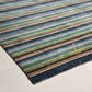 A close-up of a striped area rug with horizontal bands of navy, blue, green, beige, and brown, laid out on a light-colored floor. A gold monogram logo is visible on the lower right corner of the rug.