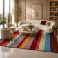 A large rectangular area rug with bold vertical rainbow stripes in red, orange, yellow, green, blue, and purple, styled in a modern living room with a curved cream sofa, matching armchair, and coffee table.