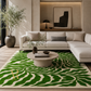 A large area rug with a bold green and cream wavy abstract pattern, styled in a modern living room setting with a cream sectional sofa, round concrete coffee table, and a potted olive tree.