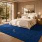 A large royal blue area rug with a gold monogram logo, styled in a modern, warm-toned bedroom next to a bed with white linens and wooden nightstands.