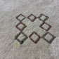 A rectangular beige area rug with a Southwestern geometric pattern in brown, gold, and black, featuring a gold monogram logo near the bottom right. The rug is shown flat against a solid white background.