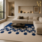A modern living room scene featuring a large cream-colored area rug with a bold blue and white circular dot pattern, styled with a beige sectional sofa and a dark wood coffee table.