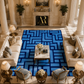 A large rectangular area rug with a bold geometric maze-like pattern in shades of blue and navy, styled in a luxurious, high-ceilinged classic living room with cream sofas, columns, and a marble fireplace mantel.