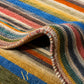 A close-up of a striped area rug with a multicolor pattern including blue, yellow, orange, green, red, and beige. The rug is shown folded to reveal its texture and underside, with a gold monogram logo in the bottom-left corner.