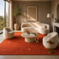 A styled interior scene with a cream curved sofa, two matching boucle armchairs, a round stone coffee table, and a large rust-red area rug. The room has warm neutral walls, wood paneling, a window with outdoor view, and decorative plants and lamps.