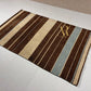 A rectangular area rug with vertical stripes in dark brown, beige, and light blue-gray, featuring a gold monogram logo in the upper right corner. The rug is displayed on a white tiled floor.