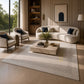 A large cream-colored area rug with a dotted border pattern is styled in a modern, sunlit living room with a curved cream sofa, two matching armchairs, and a marble coffee table.