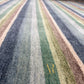 Close-up of a striped textured rug with vertical bands of navy, gray, sage green, beige, and brown, featuring a gold monogram logo near the bottom.