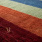 A close-up of a plush, high-pile shag rug with diagonal stripes in deep burgundy, rust orange, sage green, and blue. A gold 'AR The Artisan' logo is visible on the bottom left of the burgundy section.