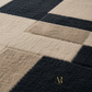 A close-up of a plush textured rug with a geometric block pattern in cream, tan, and dark charcoal gray. A gold 'R by R Design' logo is visible in the bottom right corner.