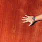 A close-up of a textured rust-orange fabric or rug surface with a person's hand and forearm resting on the right side, wearing a wristwatch. A small gold monogram logo is visible near the lower left.