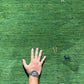 A close-up of a green textured surface resembling grass or turf, with a person's hand placed on it for scale. The hand wears a silver-toned wristwatch.