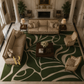 An overhead view of a luxurious living room featuring a large green and beige patterned area rug, a beige damask sofa, two matching armchairs, and a dark wood coffee table.