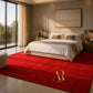 A luxurious bedroom scene featuring a bold, deep red shag area rug with a gold 'Artsisa Rumi' monogram logo in the bottom corner. The room is styled with a beige upholstered bed, white bedding, matching bedside tables, and a large window looking out onto trees.