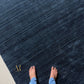 A dark navy blue area rug with a subtle horizontal textured pattern and a small gold monogram logo in the bottom right corner, shown against a plain white background.