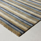 A close-up of a striped area rug with horizontal bands of beige, brown, slate blue, and charcoal gray, showing a frayed edge and a small gold embroidered logo on a light concrete floor.