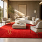 A luxurious modern living room featuring a prominent bright red area rug with a gold 'Amala Rug' logo in the bottom left corner. The room is styled with a curved white sofa, two cream armchairs, a marble coffee table, and warm natural light streaming in from a large window on the left.