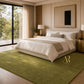A modern bedroom featuring a large olive green textured area rug on a light wood floor, styled with a cream bed, white pillows, and gold bedside tables. A gold monogram logo for 'Royal Bedding' is visible on the rug.