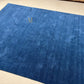 A large blue area rug with a subtle textured pattern and a small gold monogram logo near the top, laid out on a light beige tiled floor.