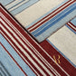 Close-up of a striped area rug with cream, light blue, and deep red bands, showing the texture and corner fold of the rug. A gold 'R' logo watermark is visible in the lower right corner.