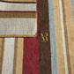 Close-up of a striped area rug with beige, brown, burgundy, and light blue vertical bands, showing a corner with a gold monogram logo reading AR and smaller text below.