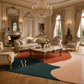 An ornate luxury living room with cream chaise lounge, matching armchairs, a marble-top coffee table, and a large abstract area rug in teal, rust orange, and blush pink. The room features classic French-style molding, a crystal chandelier, and a gold monogram logo on the rug.