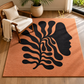 A large rectangular area rug in terracotta orange with a bold black abstract face and leaf motif, shown in a styled living room with a wooden chair, throw blanket, plant, and window.
