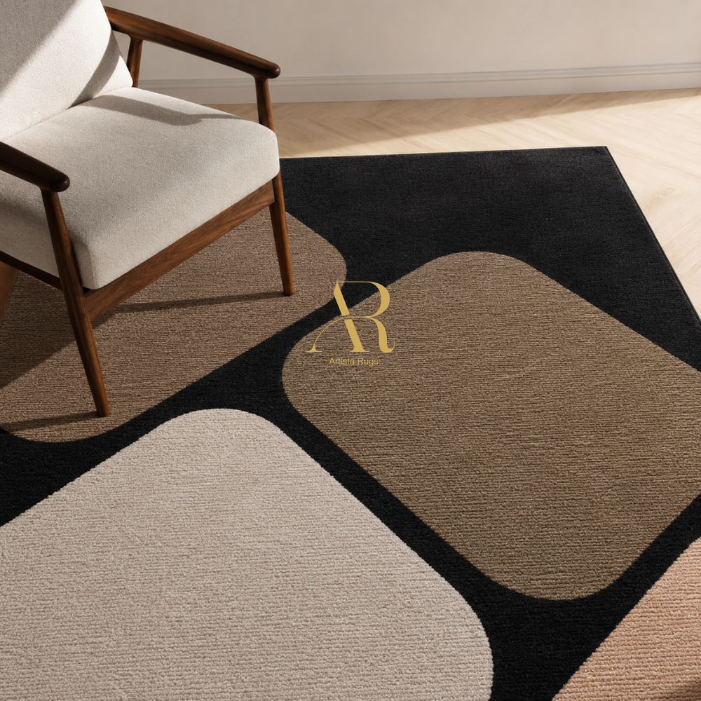 A modern area rug with a geometric abstract pattern in black, beige, taupe, and blush pink, styled in a room next to a mid-century modern armchair. A gold 'Riviera' monogram is printed on the rug.