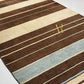 A close-up of a striped area rug with alternating wide brown and beige bands, thin cream lines, and two broad light blue stripes. A gold monogram logo is visible near the center-right.