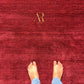 A top-down view of a person's bare feet standing on a deep red textured rug with a gold monogram logo reading 'AR' in the center.