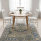 Round wooden dining table with white chairs on a patterned rug in a bright room.