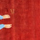 A close-up of a textured red rug with a gold 'AR' logo in the upper left, and a person's bare feet and blue jeans visible along the left edge for scale.