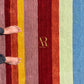 A close-up of a striped rug with vertical bands of burgundy, mustard yellow, dusty rose, deep red, orange, pale sage green, and blue. Two bare feet and black pants are visible on the left side, standing on the rug.
