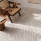 A light beige area rug with a white wavy leaf pattern, styled in a modern room with a wooden armchair and a small wooden side table.