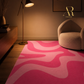A pink and magenta abstract wavy area rug styled in a modern living room setting with a wooden floor, a curved beige armchair, a floor lamp, and a wooden sideboard.