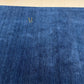 A close-up of a textured royal blue rug with a subtle gold embroidered monogram logo near the top left, set against a light beige tiled floor.
