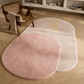 Two oval plush area rugs on a light wood herringbone floor in a styled room. One rug is dusty pink and the other is warm off-white/cream, both with a subtle gold monogram logo.