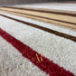 Close-up of a textured area rug with a cream base, a bold red stripe running diagonally, and stripes of brown, beige, and white in the background.
