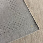 Close-up of a gray woven rug with a subtle dotted pattern, shown folded over to reveal its texture and edge against a beige floor.