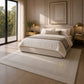 A modern bedroom scene featuring a large, light beige textured area rug with a dotted border pattern under a bed. The room has warm neutral walls, gold bedside tables and lamps, and a large window looking out onto green trees.